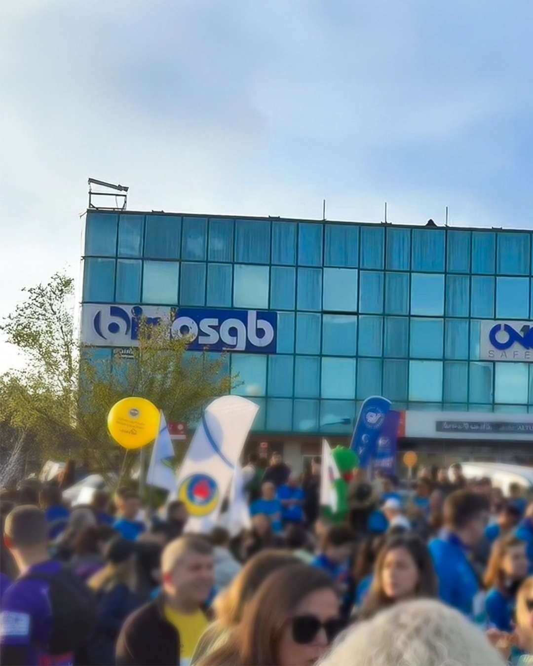 Üzerinde büyük mavi Bir OSGB tabelası bulunan kurumsal merkez binasının dış cephesi ve bina önündeki kalabalık etkinlik ortamı.